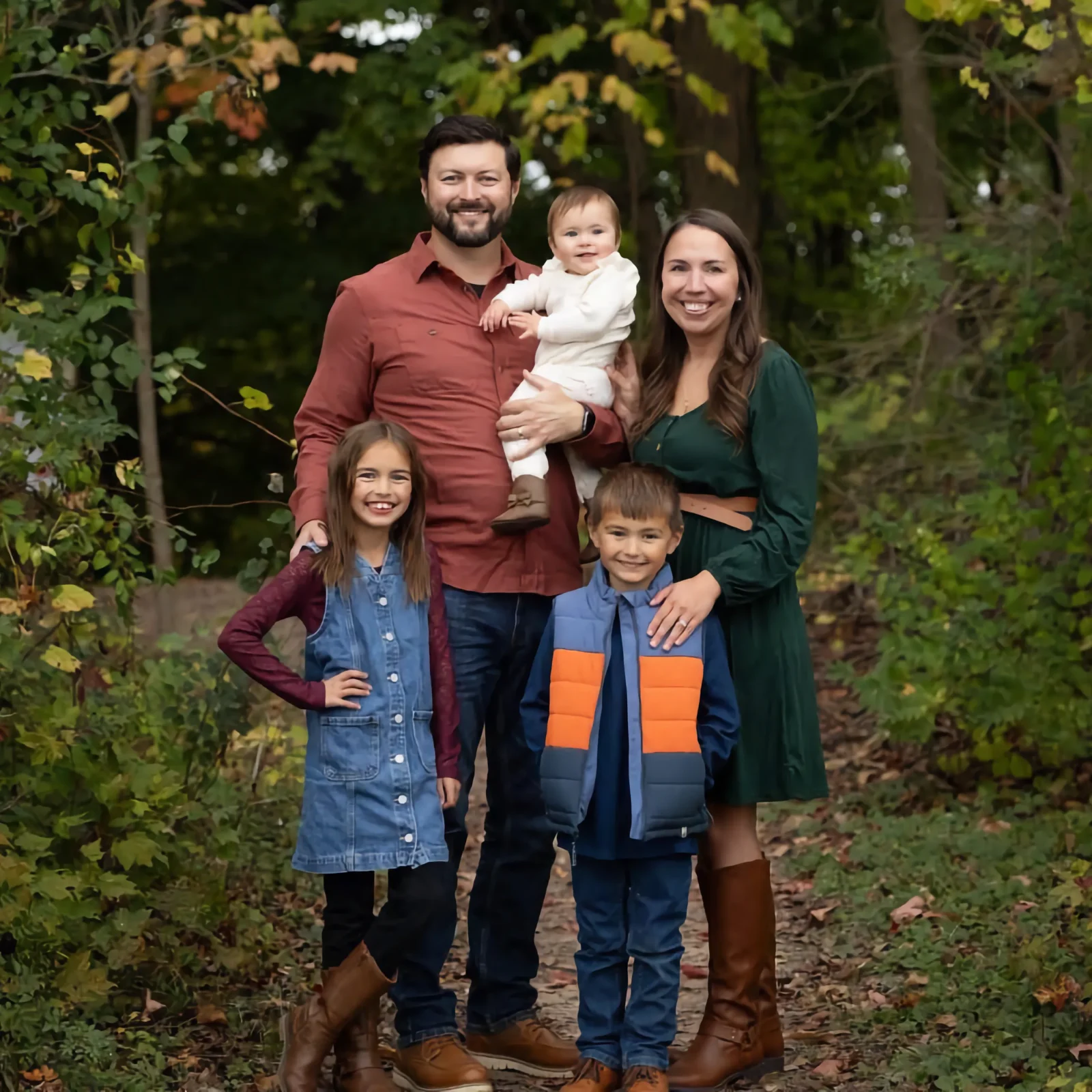 A smiling family of five stands on a wooded path: Dalton Post holds a baby next to his wife in a green dress, a girl, and a boy.