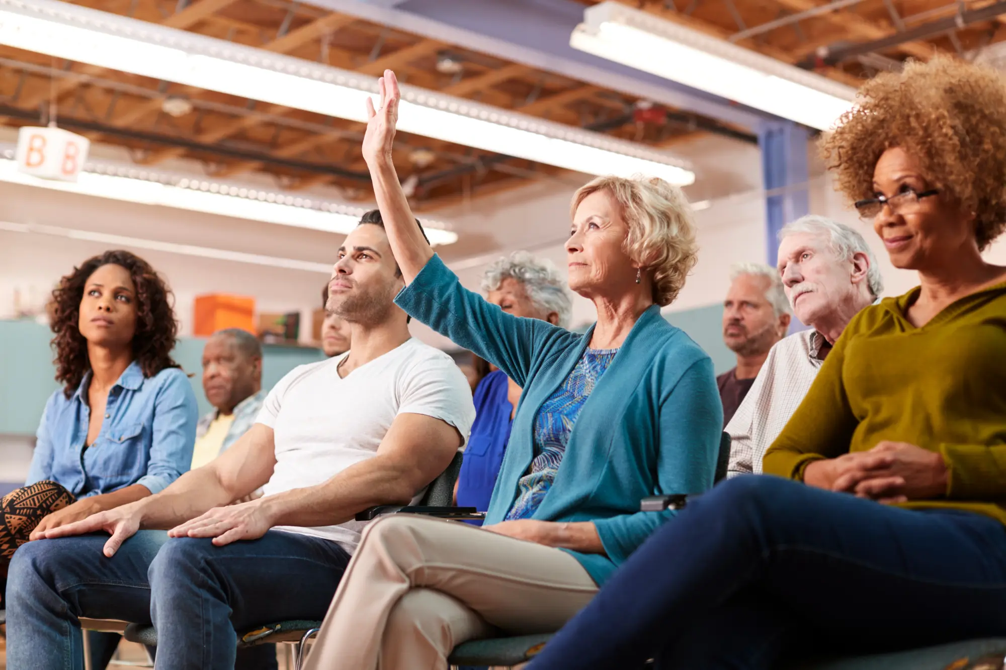 A woman with blonde hair raises her hand to speak during a community or HOA meeting while a diverse group of attendees watches attentively.
