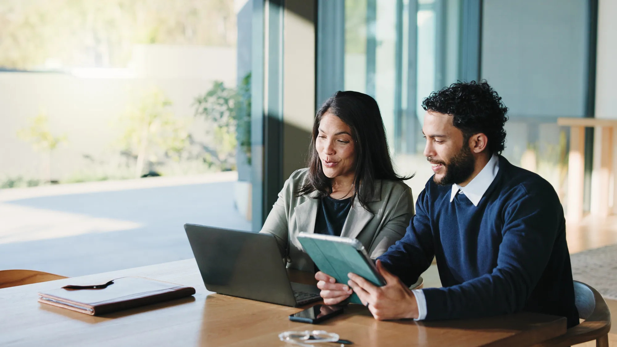 A man and a woman meet at a table in a bright office, working together on a laptop and a tablet with focused, collaborative expressions.