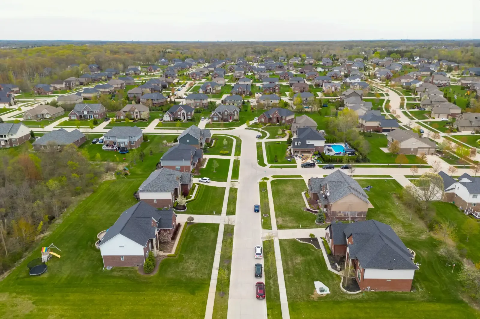 An aerial view of a large, modern suburban neighborhood featuring many single-family homes, manicured green lawns, and a quiet street leading through the center.