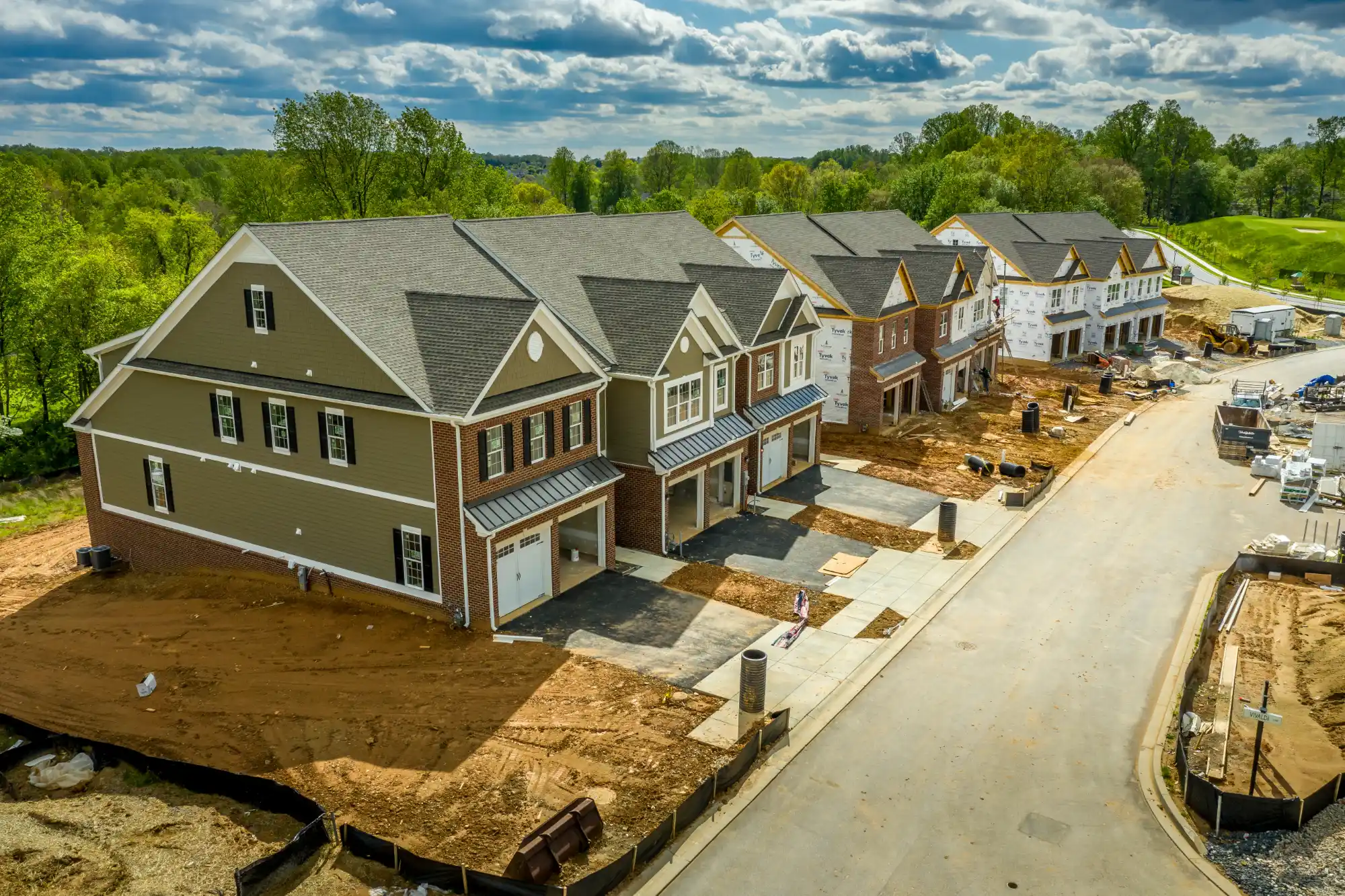An aerial view of a new residential development showing several two-story townhomes in various stages of construction lining a paved street against a green backdrop.