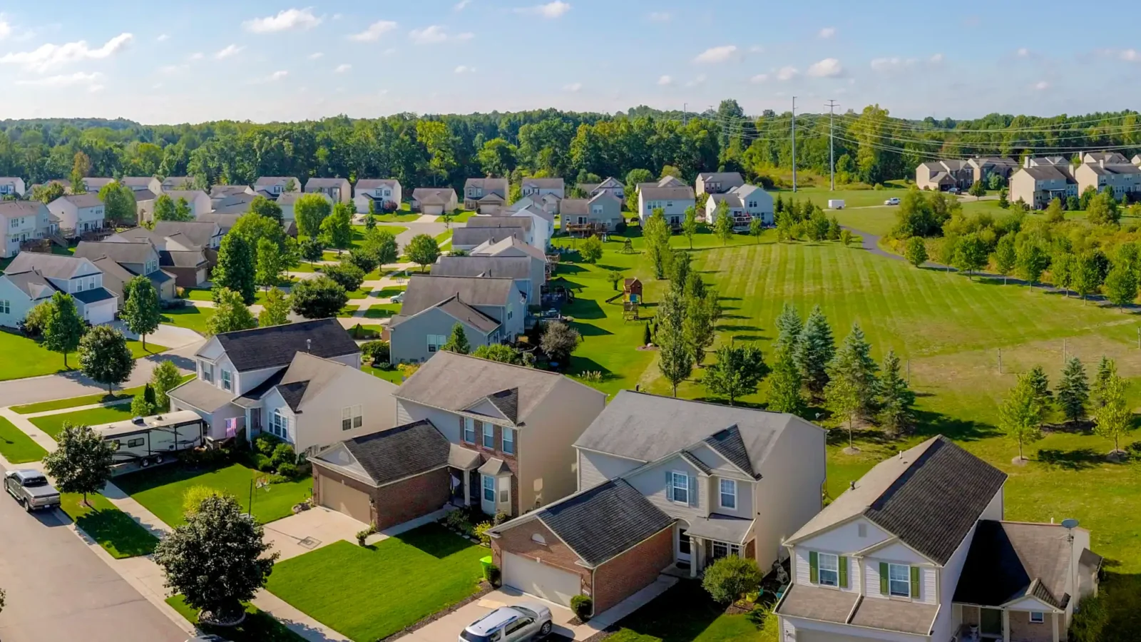 A wide aerial view of a modern suburban housing development with two-story homes and manicured lawns, bordered by a large open green space and dense woods.